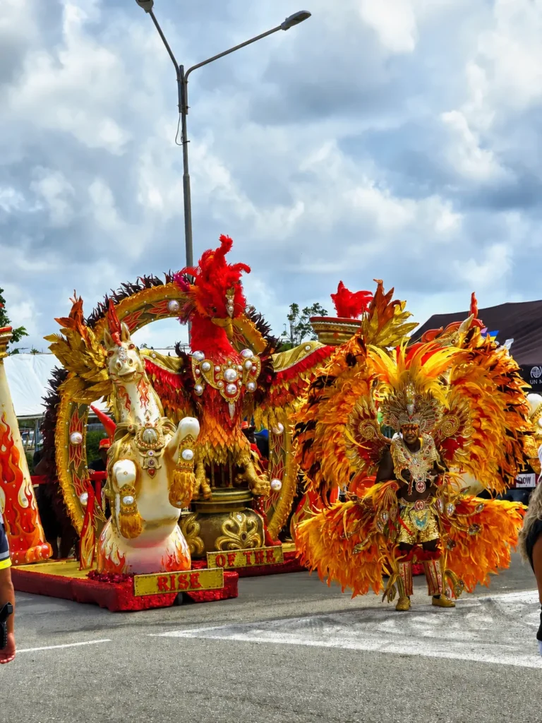 Deelnemer in een rood, oranje en goud carnavalskostuum voor een versierde praalwagen op Aruba