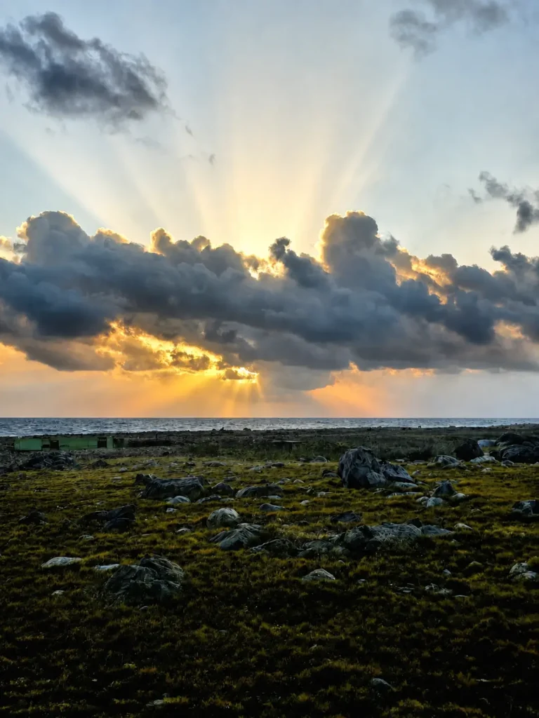 Zonnestralen breken door donkere wolken boven zee en het rotsachtige kustlandschap van Aruba